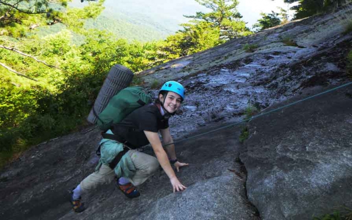 a person wearing rock climbing gear smiles at the camera as they scale a rock on an outward bound trip for lgbtq teens